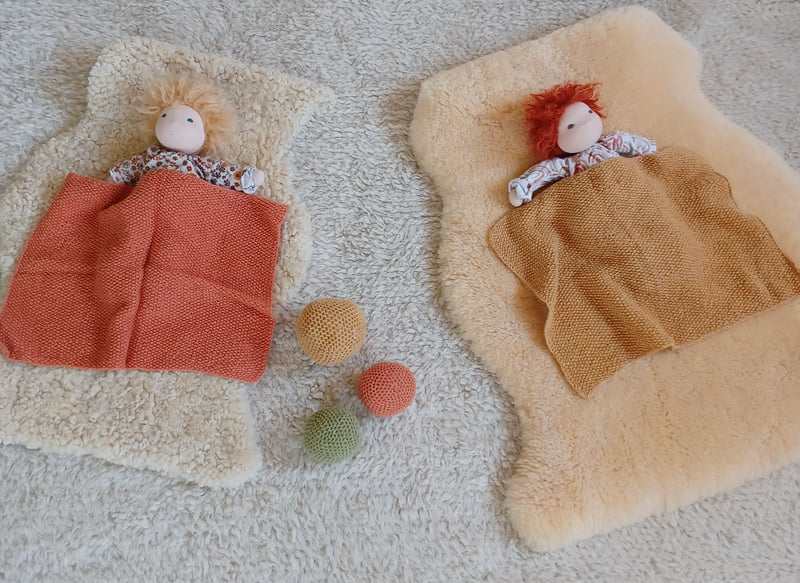 Zwei Puppen unter gestrickten Decken auf Lammfellen, daneben Filzkugeln